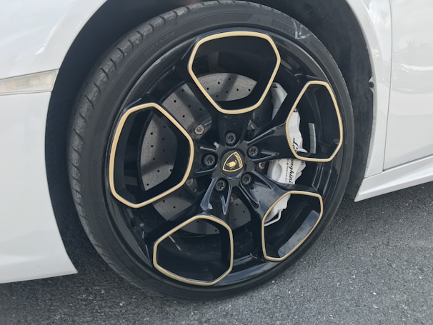 A close-up view of a luxury car wheel featuring a glossy black rim with gold accents in a geometric pattern. The tire has visible tread, and the car is white. The brake caliper displays branding.