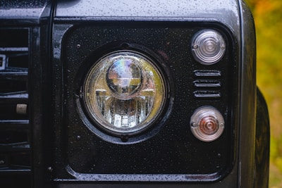 A close-up view of a vehicle's front headlight, surrounded by a black frame with raindrops on the surface. Two additional smaller lights are visible on the right, with one appearing slightly orange. The background is blurred with a hint of green and yellow.