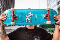 A person holding a turquoise skateboard in front of their face. The skateboard has red trucks and the word 'Fly' printed on it along with the phrase 'High Rollin'. The person has tattoos on their arms and is wearing a black shirt and a beaded necklace. The background features an urban setting with modern buildings and greenery.