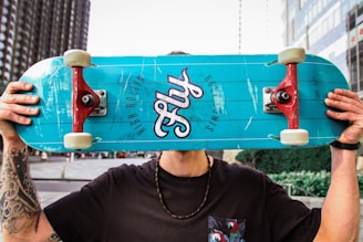 A person holding a turquoise skateboard in front of their face. The skateboard has red trucks and the word 'Fly' printed on it along with the phrase 'High Rollin'. The person has tattoos on their arms and is wearing a black shirt and a beaded necklace. The background features an urban setting with modern buildings and greenery.