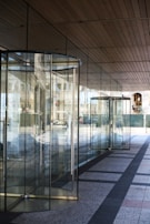 A series of glass revolving doors reflecting the outside street scene, with a modern design and metal frames. The clean architectural lines of the building and its ceiling panels are visible with reflections of the street creating a dynamic interplay of glass and light.