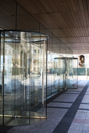 A series of glass revolving doors reflecting the outside street scene, with a modern design and metal frames. The clean architectural lines of the building and its ceiling panels are visible with reflections of the street creating a dynamic interplay of glass and light.