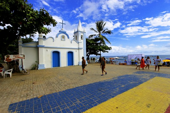 A small white church with blue accents and a cross on top sits near a beach under a bright blue sky, surrounded by palm trees. People are walking on the paved area in front of the church, and a few are seated nearby under umbrellas. To the side, there is a sign pointing to Pra&ccedil;a S&atilde;o Francisco, and boats are visible in the ocean in the background.