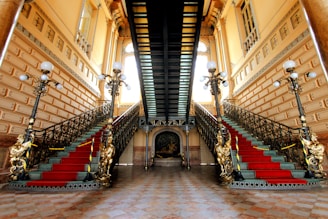 An opulent interior featuring a grand double staircase adorned with ornate railings and red carpet runners. The setting is illuminated by vintage-style floor lamps with multiple bulbs, and the walls are decorated with intricate moldings and large windows.