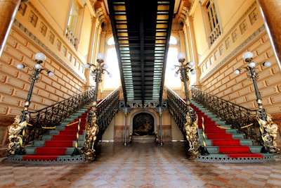 An opulent interior featuring a grand double staircase adorned with ornate railings and red carpet runners. The setting is illuminated by vintage-style floor lamps with multiple bulbs, and the walls are decorated with intricate moldings and large windows.