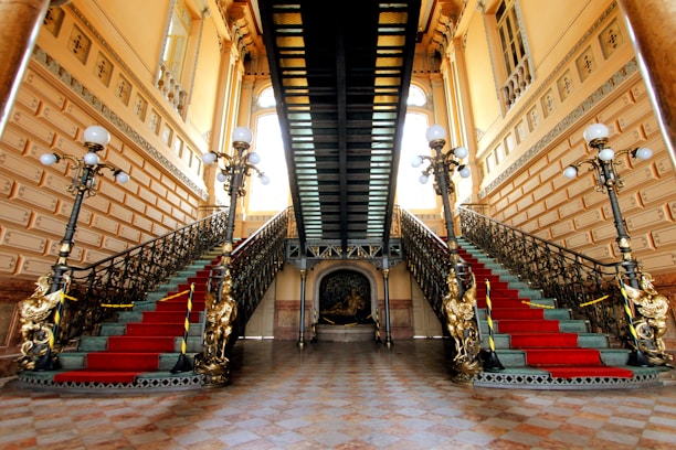 An opulent interior featuring a grand double staircase adorned with ornate railings and red carpet runners. The setting is illuminated by vintage-style floor lamps with multiple bulbs, and the walls are decorated with intricate moldings and large windows.