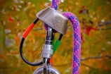 A close-up view of climbing equipment, featuring a metallic belay device attached to a sturdy rope and a carabiner. The background is blurred, with autumn-colored leaves visible.