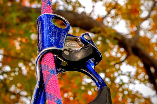 A close-up of a blue belaying or climbing device attached to a multicolored rope. The background features a blurred view of autumn foliage with orange and green leaves.