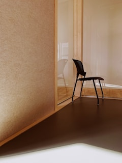 A minimalistic interior space features a corner with a single black chair positioned on a wooden floor. The walls are made of light-colored wood and glass, creating a modern and clean design. Sunlight casts soft shadows on the floor, enhancing the warm tones.