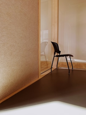 A minimalistic interior space features a corner with a single black chair positioned on a wooden floor. The walls are made of light-colored wood and glass, creating a modern and clean design. Sunlight casts soft shadows on the floor, enhancing the warm tones.