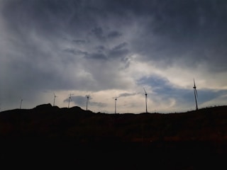 Rows of tall, white wind turbines standing against a dramatic cloudy sky over rolling hills.