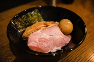 A bowl filled with a large slice of pink, cooked pork, a marinated egg with a shiny brown surface, and a side of dark leafy greens, set on a wooden table.