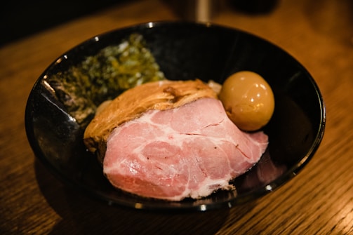 A bowl filled with a large slice of pink, cooked pork, a marinated egg with a shiny brown surface, and a side of dark leafy greens, set on a wooden table.