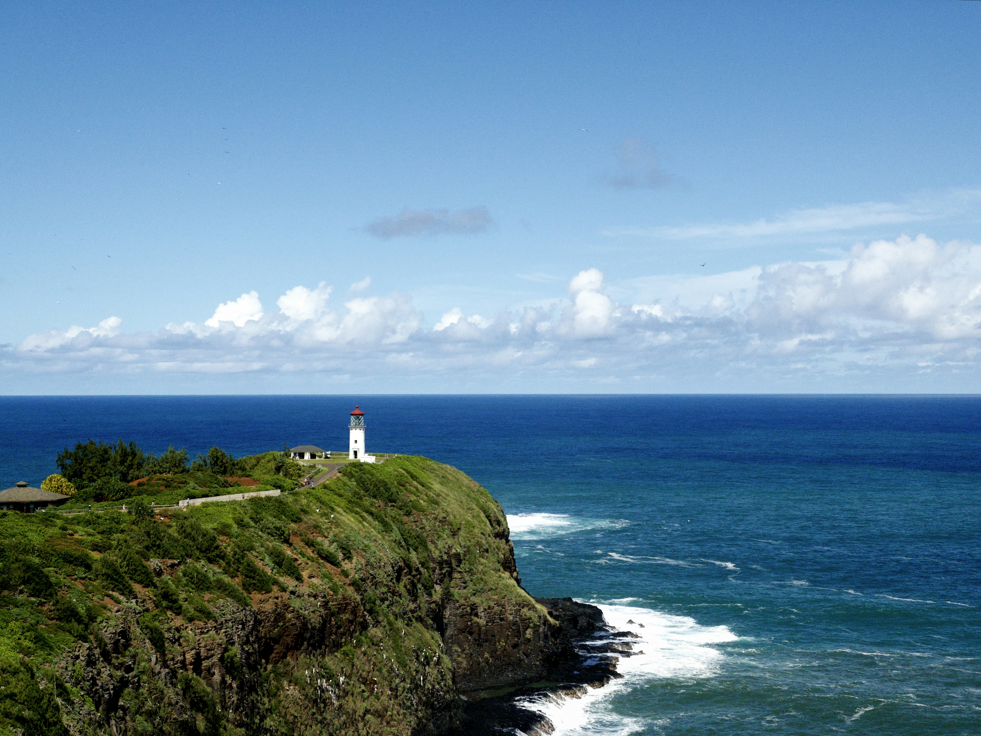 A photo of the Kilauea Lighthouse on Kauai, Hawaii. The white lighthouse with a red roof stands on a cliff overlooking the blue Pacific Ocean. Lush green vegetation and potentially other parts of the Kilauea Point National Wildlife Refuge are visible in the background. [Photo by Jason Weingardt on Unsplash]