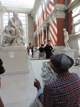 An artist is sketching a sculpture inside a sunlit gallery with high ceilings and large windows. The sculpture depicts a group of intertwined figures, and the artist appears focused on capturing its details. Other visitors are observing various artworks in the lively museum environment.