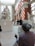 An artist is sketching a sculpture inside a sunlit gallery with high ceilings and large windows. The sculpture depicts a group of intertwined figures, and the artist appears focused on capturing its details. Other visitors are observing various artworks in the lively museum environment.
