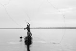 A person casts a fishing net over a calm body of water from a wooden boat. The scene is tranquil with a subtle reflection on the water's surface, and the horizon is visible in the distance under a cloudy sky.