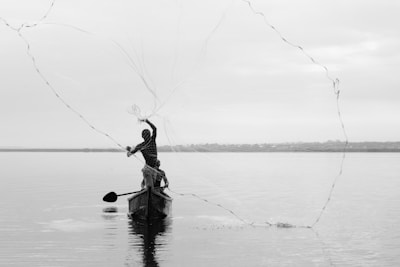 A person casts a fishing net over a calm body of water from a wooden boat. The scene is tranquil with a subtle reflection on the water's surface, and the horizon is visible in the distance under a cloudy sky.