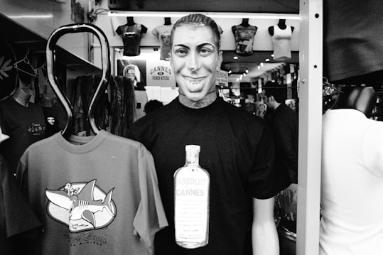 A mannequin wearing a T-shirt with a graphic of a bottle labeled 'Absolut Cannes' is displayed prominently in a shop. Next to it, another T-shirt features a cartoonish shark. The shop is filled with various other merchandise, including clothes hanging on the walls. The setting appears to be a busy retail space.