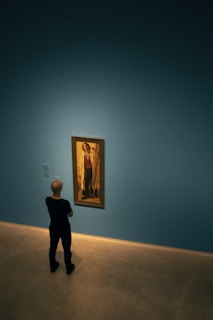 A person standing in a minimalistic gallery room, observing a framed painting on a blue wall. The painting is displayed in a well-lit area, creating a contrast with the overall dimness of the room.