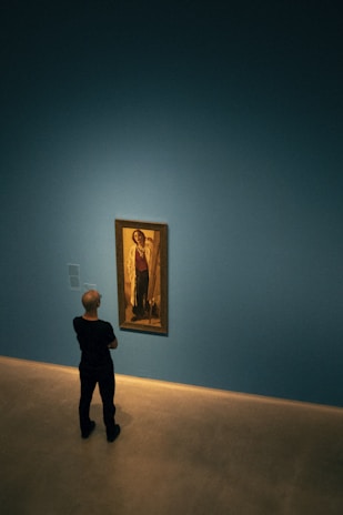 A person standing in a minimalistic gallery room, observing a framed painting on a blue wall. The painting is displayed in a well-lit area, creating a contrast with the overall dimness of the room.