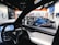 The interior view of a Tesla car focusing on the steering wheel and digital dashboard, with a blue Tesla car visible through the driver's side window. Surrounding the vehicles, posters and screens showcase various Tesla features in a showroom environment with bright lighting.