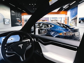 The interior view of a Tesla car focusing on the steering wheel and digital dashboard, with a blue Tesla car visible through the driver's side window. Surrounding the vehicles, posters and screens showcase various Tesla features in a showroom environment with bright lighting.
