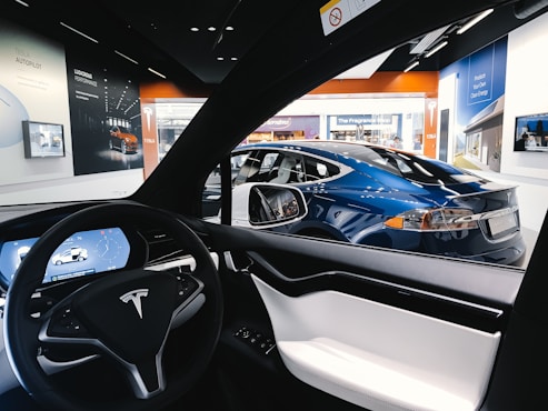 The interior view of a Tesla car focusing on the steering wheel and digital dashboard, with a blue Tesla car visible through the driver's side window. Surrounding the vehicles, posters and screens showcase various Tesla features in a showroom environment with bright lighting.