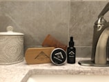 A bathroom countertop featuring a ceramic jar with a decorative pattern, a wooden beard brush, a beard balm container, a wooden comb, and a black bottle of beard oil. The arrangement is set against a tiled wall, and a stainless steel tap is visible on the right side.