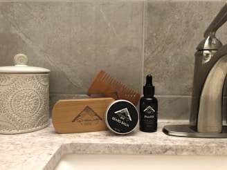 A bathroom countertop featuring a ceramic jar with a decorative pattern, a wooden beard brush, a beard balm container, a wooden comb, and a black bottle of beard oil. The arrangement is set against a tiled wall, and a stainless steel tap is visible on the right side.