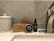 A bathroom countertop featuring a ceramic jar with a decorative pattern, a wooden beard brush, a beard balm container, a wooden comb, and a black bottle of beard oil. The arrangement is set against a tiled wall, and a stainless steel tap is visible on the right side.