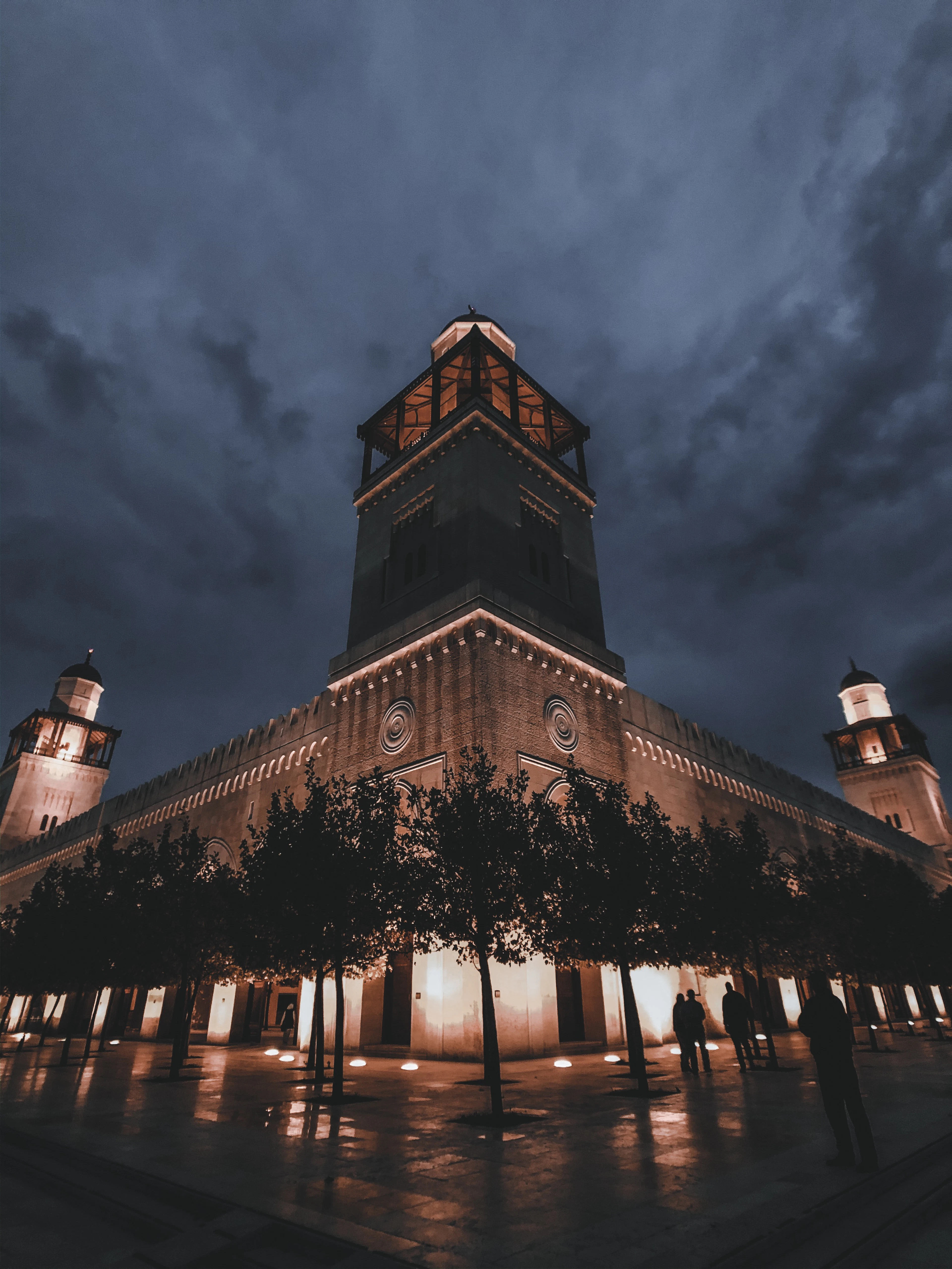 A floodlit stone landmark with corner minarets and a lantern-topped tower in downtown Amman at night, its arcade lights reflected on wet paving with only a few people crossing the empty square.