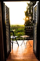 Two chairs and a small table are placed on a tiled balcony, framed by open dark shutters. The balcony overlooks a lush green landscape with trees and a hazy horizon, while the sun sets, casting a warm golden glow.