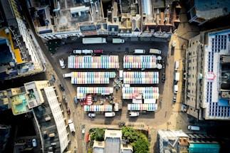 An aerial view of an urban area with a market featuring several rows of stalls. The stalls have colorful striped roofs in blue, green, yellow, and red. Surrounding buildings create a rectangular enclosure around the market. The street is populated with parked cars and small delivery trucks.