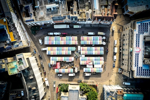 An aerial view of an urban area with a market featuring several rows of stalls. The stalls have colorful striped roofs in blue, green, yellow, and red. Surrounding buildings create a rectangular enclosure around the market. The street is populated with parked cars and small delivery trucks.