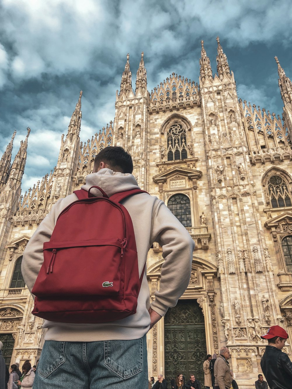 昼間、白と青の空の下、イタリアのミラノ大聖堂の近くに立つ灰色のジャケットを着て赤いリュックサックを背負った男性