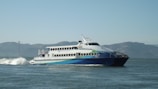 A large passenger ferry moving through the water with visible waves behind it. The ferry is white with blue and green accents and the words 'San Francisco Bay Ferry' on the side. In the background, there are hills and a faint view of what appears to be the Golden Gate Bridge under a clear sky.