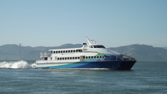 A large passenger ferry moving through the water with visible waves behind it. The ferry is white with blue and green accents and the words 'San Francisco Bay Ferry' on the side. In the background, there are hills and a faint view of what appears to be the Golden Gate Bridge under a clear sky.