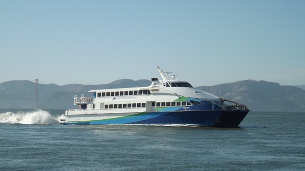 A large passenger ferry moving through the water with visible waves behind it. The ferry is white with blue and green accents and the words 'San Francisco Bay Ferry' on the side. In the background, there are hills and a faint view of what appears to be the Golden Gate Bridge under a clear sky.