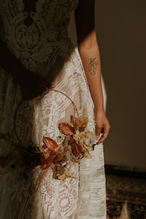 A close-up view of a person wearing an intricate lace dress holding a hoop decorated with dried flowers and leaves. The person's arm features a tattoo of a geometric-style anatomical heart. Soft lighting creates a warm and intimate atmosphere.