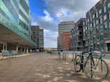 A modern urban setting features a row of contemporary buildings with glass facades on one side and dark brick designs on the other. The scene displays a clean, spacious pedestrian area with metal benches and bicycles parked along the side. The sky above is partly cloudy, casting diffused light over the area.