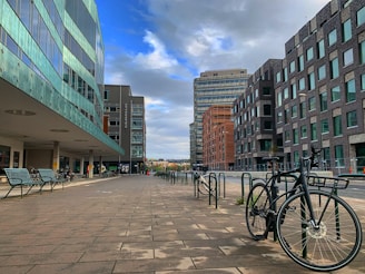 A modern urban setting features a row of contemporary buildings with glass facades on one side and dark brick designs on the other. The scene displays a clean, spacious pedestrian area with metal benches and bicycles parked along the side. The sky above is partly cloudy, casting diffused light over the area.