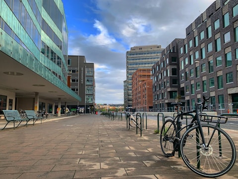 A modern urban setting features a row of contemporary buildings with glass facades on one side and dark brick designs on the other. The scene displays a clean, spacious pedestrian area with metal benches and bicycles parked along the side. The sky above is partly cloudy, casting diffused light over the area.