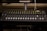 A professional audio mixing console with multiple faders, buttons, and knobs, used for adjusting sound levels and effects. It is placed on a desk with some electronic equipment visible in the background.