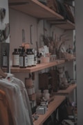 A wooden shelf displays a variety of small bottles, jars, and packages, likely personal care or aromatic products. There are also dried plants and decorative items arranged among the products, creating a rustic and cozy atmosphere. A hanging garment can be seen in the foreground, adding to the casual shopping environment.