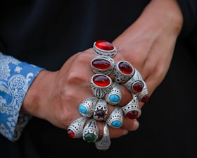 A person's hands are adorned with multiple ornate silver rings, each featuring large, polished stones in vibrant red, blue, and green colors. The intricate metalwork of the rings displays detailed patterns and craftsmanship. The background is dark, contrasting with the blue patterned fabric of the shirt sleeve.