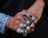 A person's hands are adorned with multiple ornate silver rings, each featuring large, polished stones in vibrant red, blue, and green colors. The intricate metalwork of the rings displays detailed patterns and craftsmanship. The background is dark, contrasting with the blue patterned fabric of the shirt sleeve.