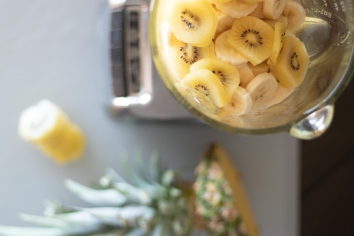 A glass container filled with sliced bananas and kiwifruit is placed on a kitchen scale. A whole pineapple is partially visible at the bottom, and a blurred jar of what appears to be yellow juice or smoothie is on the left side of the image.