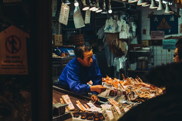 A vendor in a blue shirt interacts with a customer at a bustling market stall, surrounded by a variety of packaged and fresh seafood. The stall is illuminated by hanging lights, and various signs with text in a foreign language are visible. The environment appears crowded and lively, with assorted goods displayed prominently on the counter.