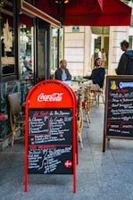 Outdoor seating area of a café with two men seated at a table, and another person standing nearby. The scene includes a red Coca-Cola branded menu board displaying breakfast options in both French and English. A secondary menu board is present on the right, listing various dishes. The café has a classic Parisian look with wicker chairs and a red awning.